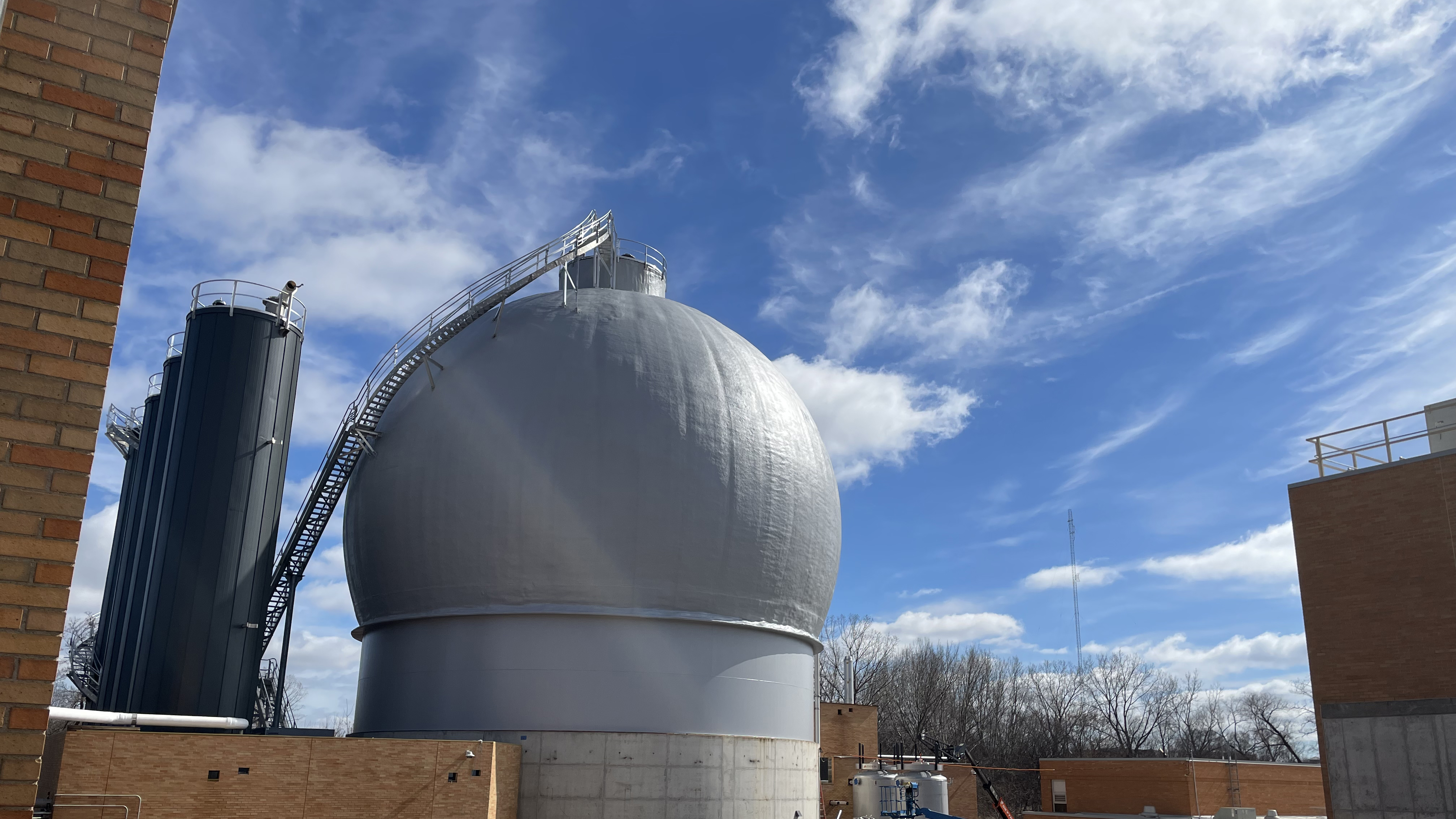 Grey ball digester near three dark grey tanks and yellow brick buildings